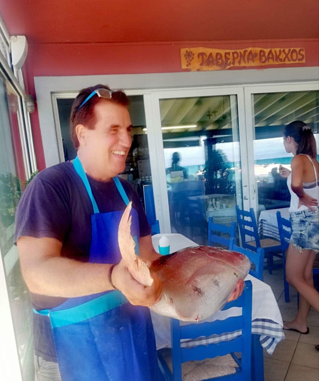 Dimitris, founder of Bacchus, laughing with a fresh red snapper under the Taverna Bakchos sign