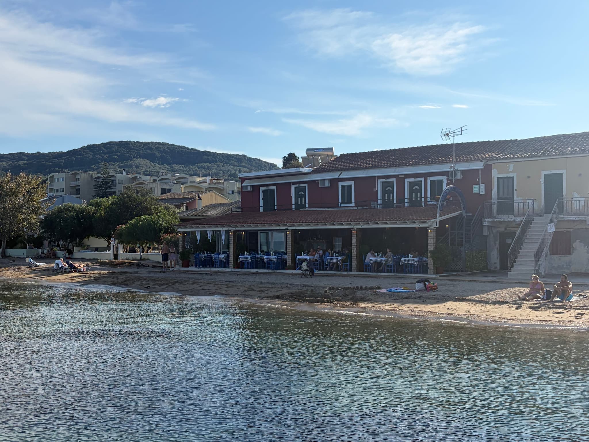 Bacchus taverna seen from the water — the red house, blue chairs under the pergola, guests at lunch, the beach in front